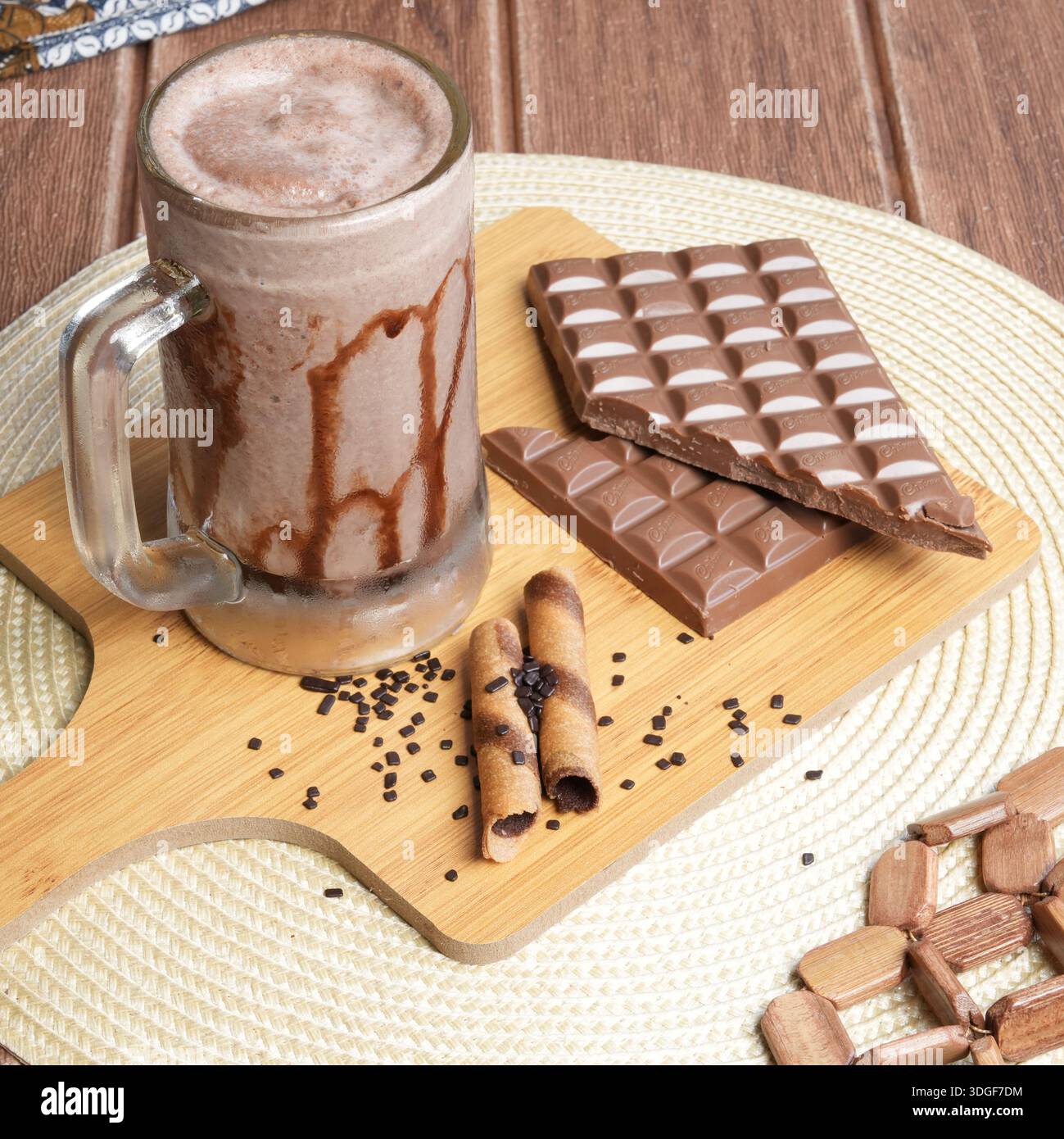 Bevanda al cioccolato ghiacciata con filo di sciroppo, abbinata a barrette di cioccolato al latte e wafer roll su un asse di legno. Foto Stock