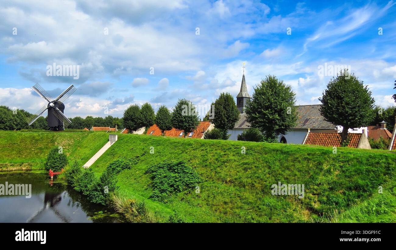 Un pittoresco paesaggio di Bourtange caratterizzato da un tradizionale mulino a vento nero, una guglia da chiesa e case storiche con tetti arancioni. Foto Stock