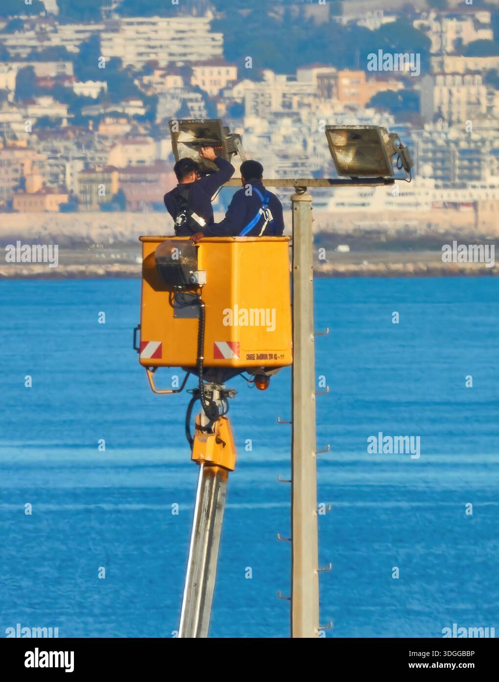 Due lavoratori in Un sollevatore a benna giallo che ispezionano Un palo luminoso sul mare con sfondo cittadino Foto Stock