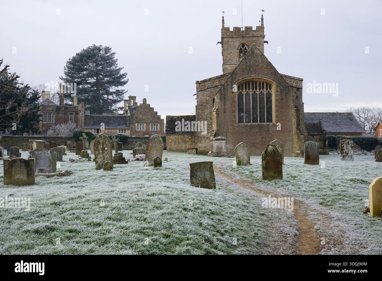 Frost copre un cimitero di un villaggio a Combe, Oxfordshire, Regno Unito, in inverno, con lapidi, un sentiero e la storica chiesa in pietra. Foto Stock