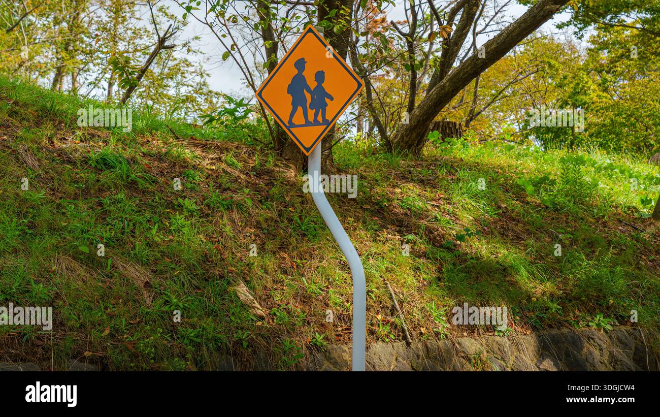 Sakata, Yamagata, Giappone - 5 ottobre 2024, vista panoramica del cartello stradale arancione "attenzione: Attraversamento dei bambini", su un pilastro curvo tra alberi, senza persone Foto Stock