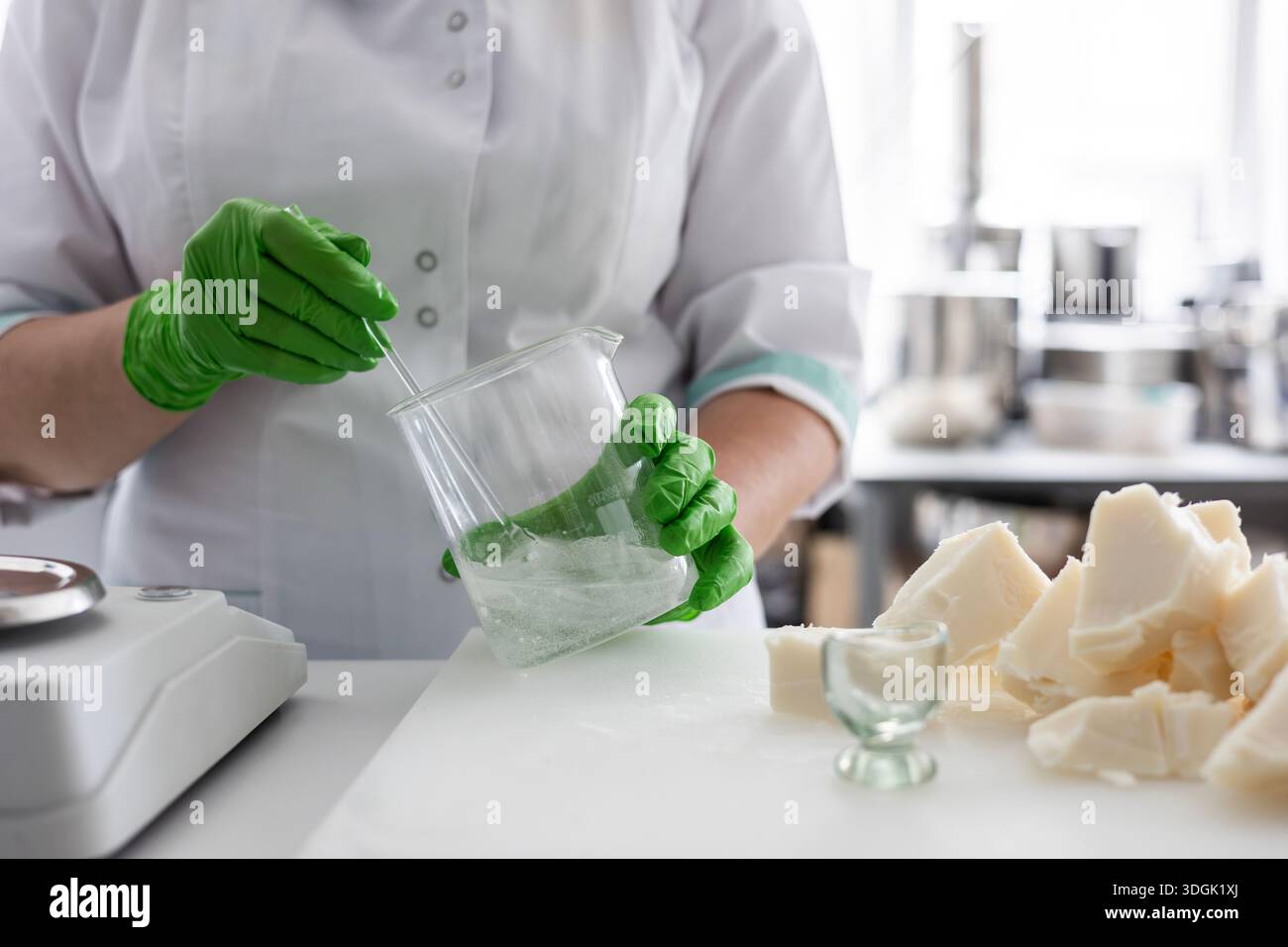 Un tecnico di laboratorio con rivestimento bianco e guanti in nitrile verde mescola un liquido in un becher. Сraft la produzione di cosmetici biologici implica la pesatura i. Foto Stock