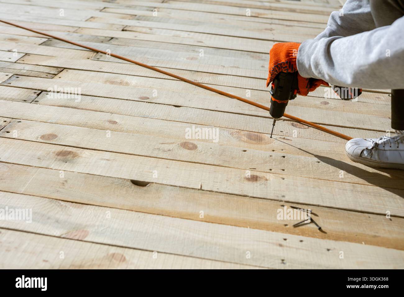 Un lavoratore con guanti arancioni utilizza un trapano cordless per inserire una vite, pericolosamente vicino a una prolunga sotto tensione. Una pratica di lavoro non sicura crea una r Foto Stock