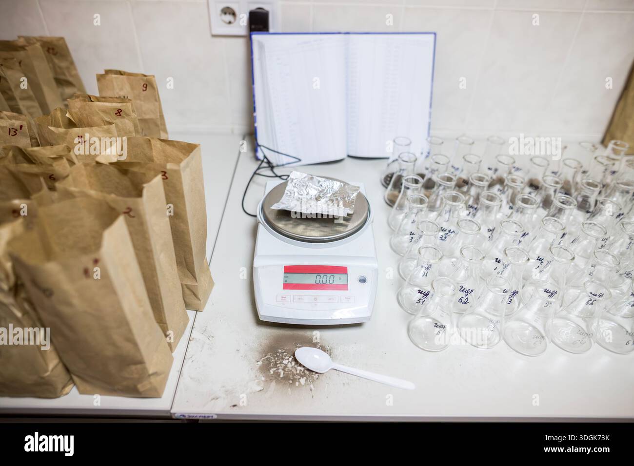 Bilancia analitica elettronica si trova sul tavolo bianco vicino ai sacchetti di carta marrone e alle matracci di vetro con campioni di terreno pronti per l'uso chimico-agrochimico Foto Stock