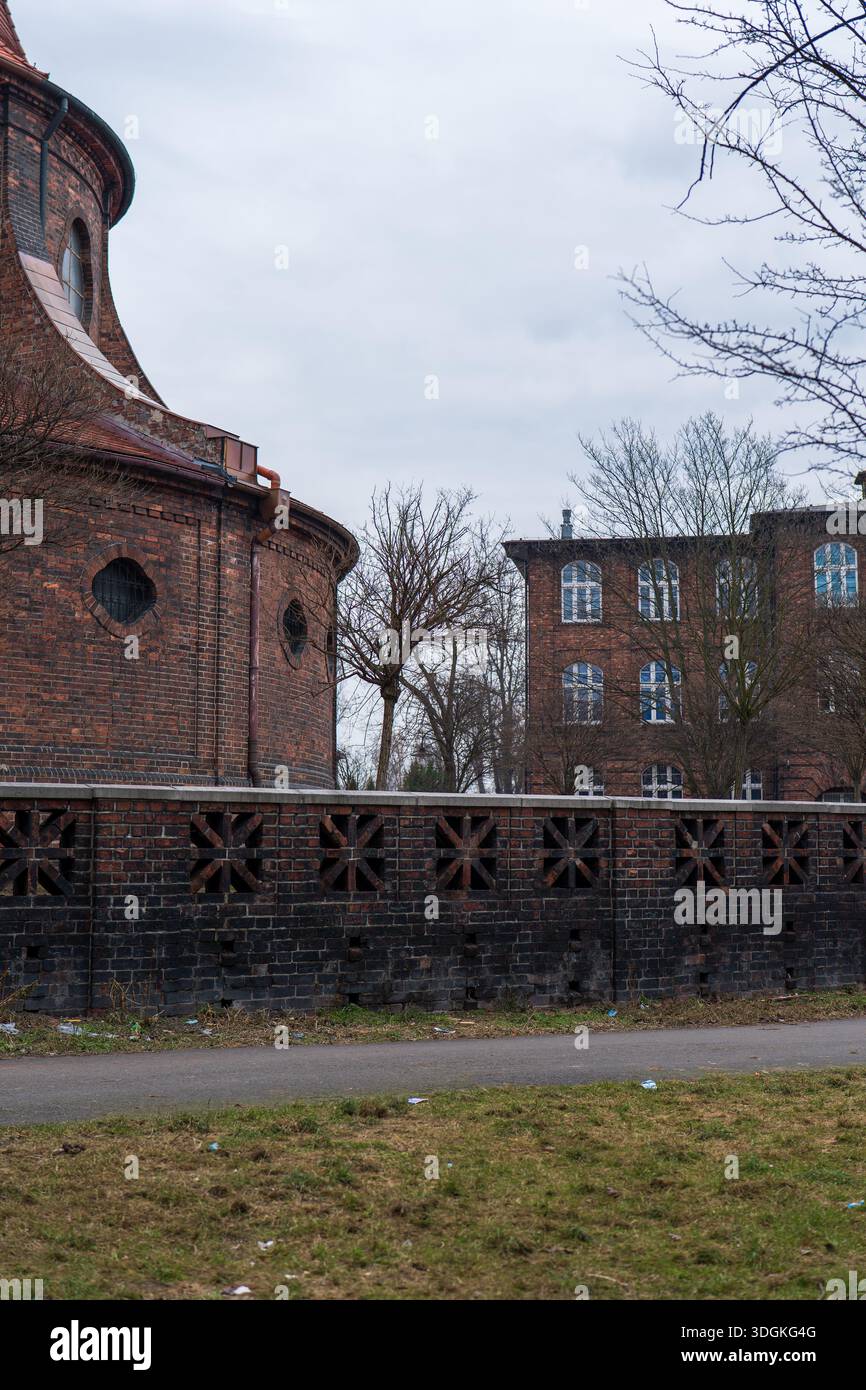 Una chiesa in mattoni con abside arrotondata si affaccia su una tenuta in mattoni a Katowice. Alberi senza fronzoli, sentiero stretto, recinzione in mattoni con aperture a stella e erba disseminata A. Foto Stock