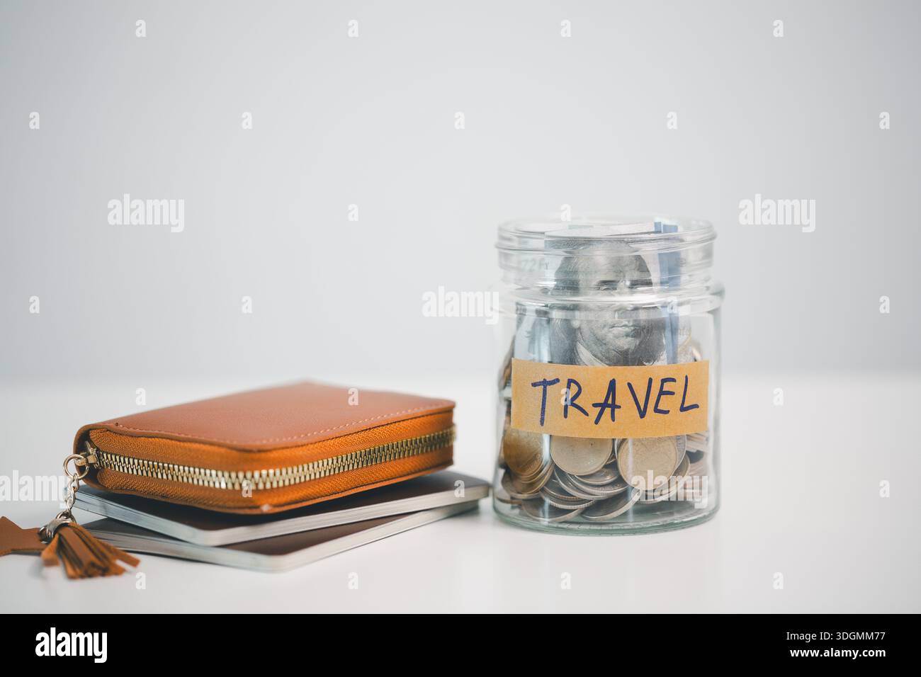 Vasetto in vetro etichettato TRAVEL pieno di monete e contanti accanto a un portafoglio in pelle e quaderni su sfondo bianco, simbolo di risparmio per le vacanze, PL Foto Stock
