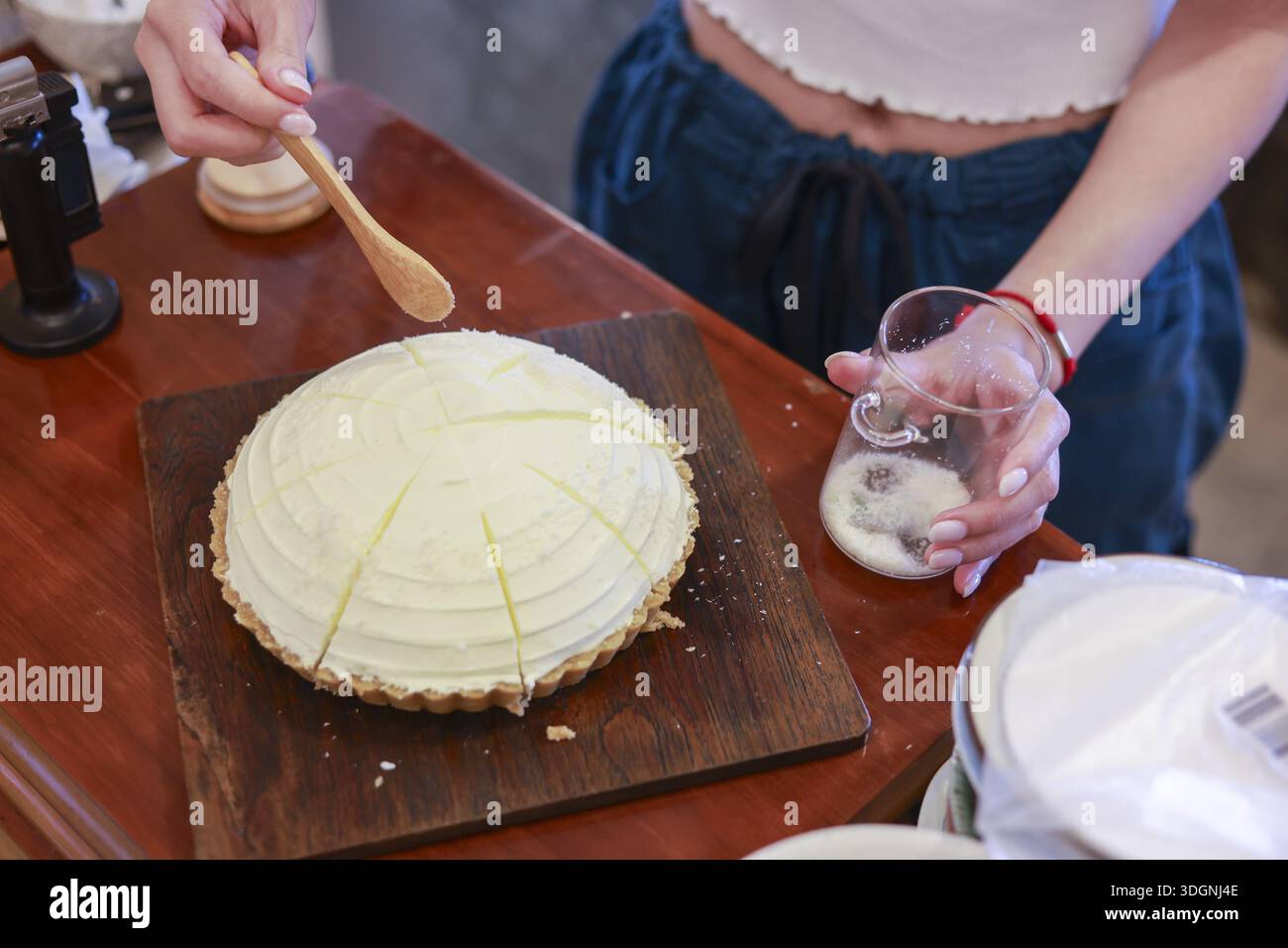 Primo piano donna che tiene a mano un cucchiaio di legno che spalma la crema dolce bianca su una deliziosa torta di cocco fatta in casa su un vassoio di legno che si prepara a servire Foto Stock