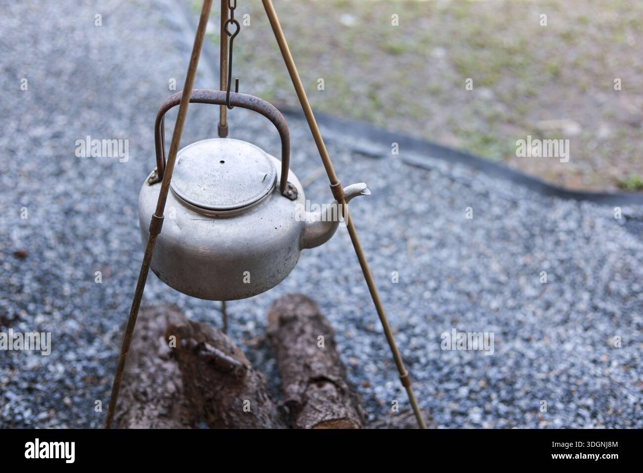 Ambiente di campeggio all'aperto con vecchio bollitore in metallo rustico appeso su treppiede sopra i tronchi di falò su terreno di ghiaia che mostra un'atmosfera tranquilla e tranquilla di atmos Foto Stock