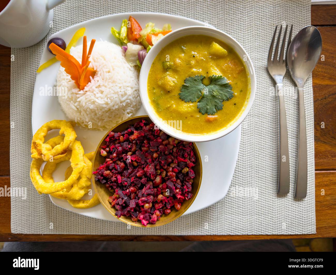 Un pasto colorato su un piatto bianco: Riso al vapore, curry vegetale con coriandolo, insalata di barbabietole, anelli di cipolla croccanti e un lato del tè su un tavolo di legno l'11 gennaio 2026 a Lindenfels im Odenwald, Germania. Lindenfels è famosa per il trattamento ayurvedico e per il castello di Lindenfels. Foto Stock