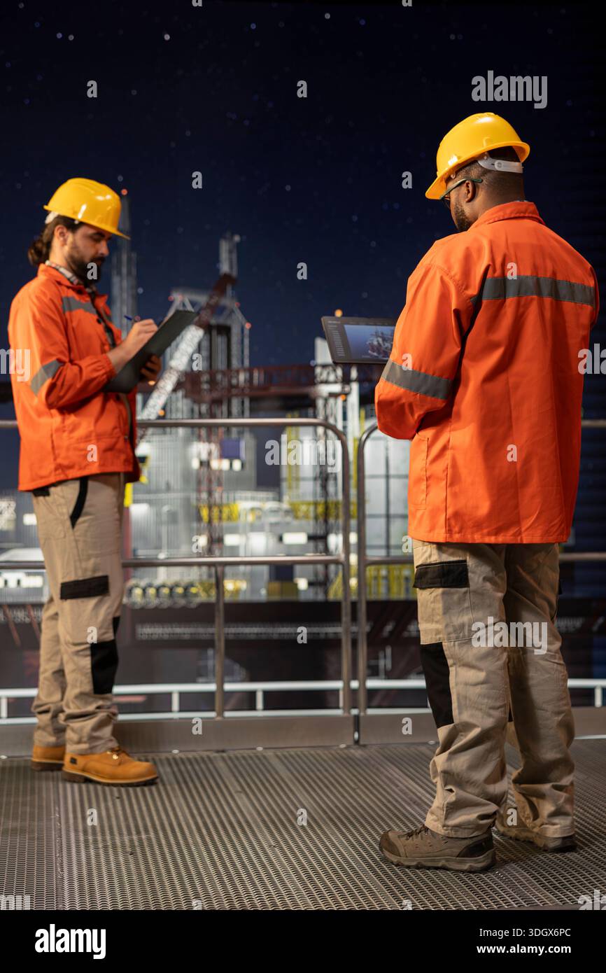 Equipaggio della piattaforma offshore che utilizza un tablet per supervisionare le operazioni di perforazione durante il turno di notte. I membri del personale addetto alla perforazione delle chiatte che esaminano i dati dei sensori sul dispositivo per garantire una produzione continua Foto Stock