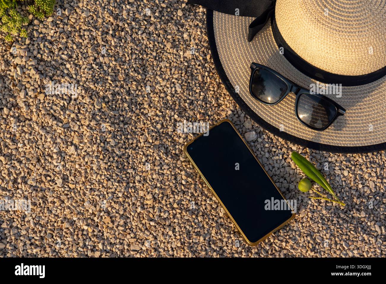 Un piatto piatto piatto degli indispensabili estivi con cappello di paglia e smartphone su piccole pietre sotto la luce del sole. Concetto di pianificazione delle vacanze, blog di viaggio, mobi Foto Stock