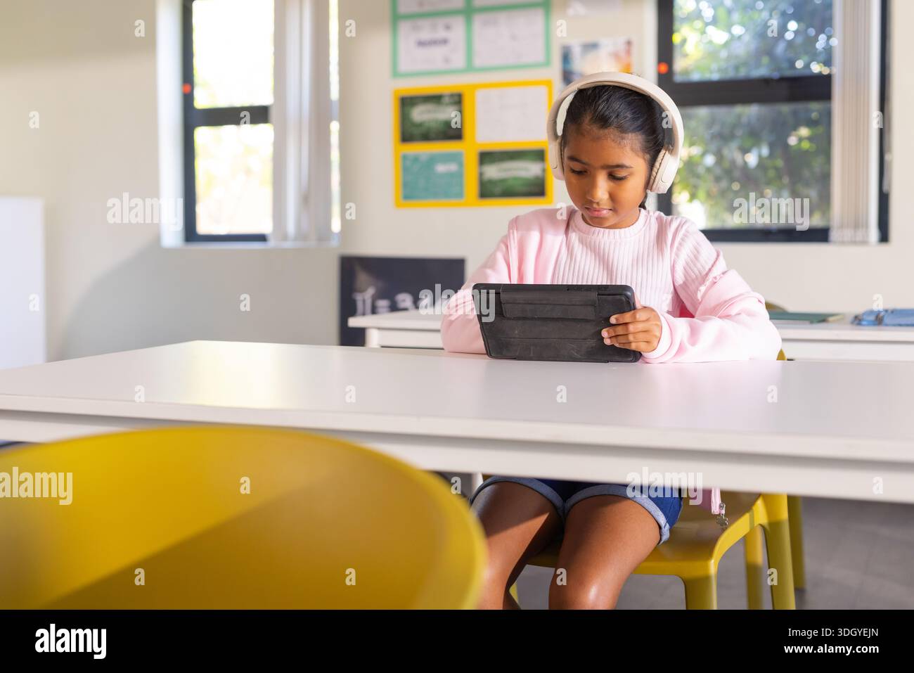 Ragazza afroamericana preadolescente seduta alla scrivania in classe con cuffie che reggono un tablet nero Foto Stock