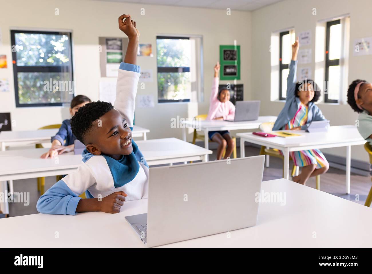 Bambini diversi seduti in classe usando un computer portatile e alzando le mani alle scrivanie bianche con poster Foto Stock