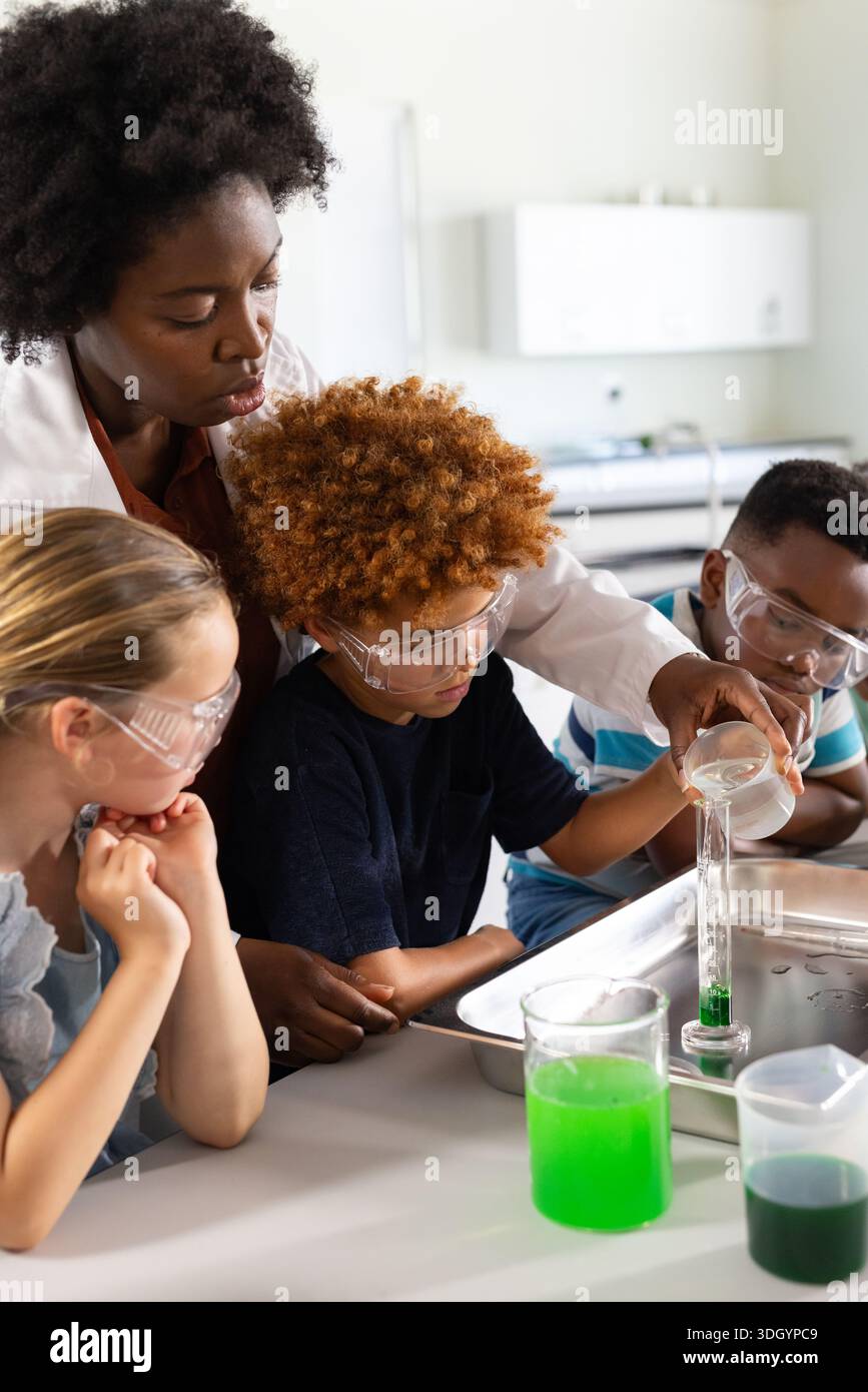 Insegnante afroamericano che guida i bambini che versano nel cilindro indossando un camice da laboratorio e occhiali di protezione Foto Stock