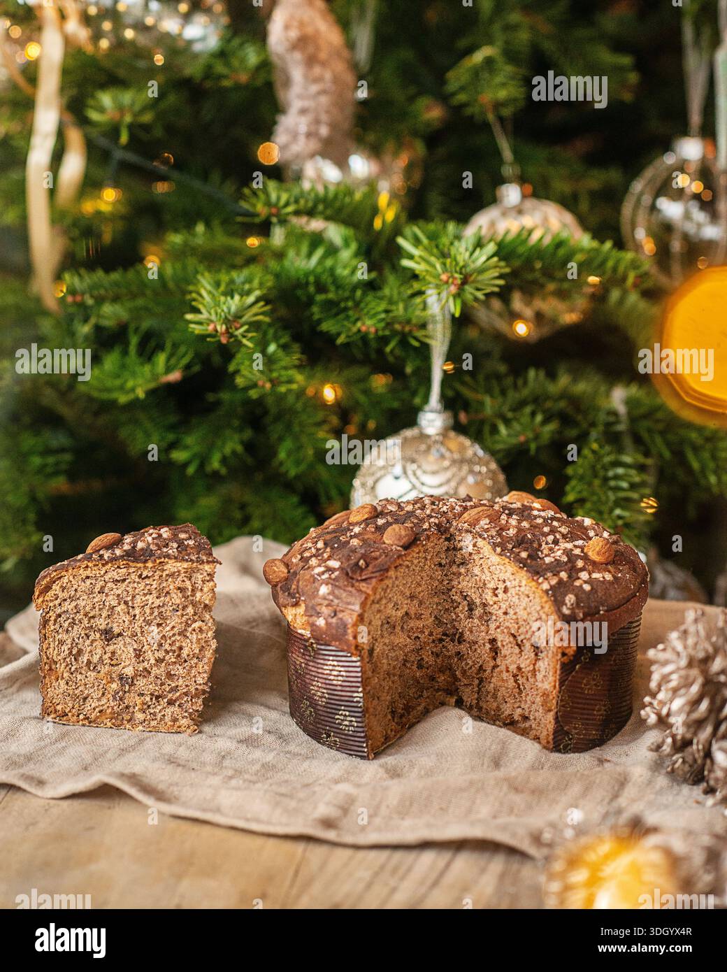 Fetta di panettone tradizionale con mandorle e zucchero perlato contro un albero di Natale decorato. Foto Stock