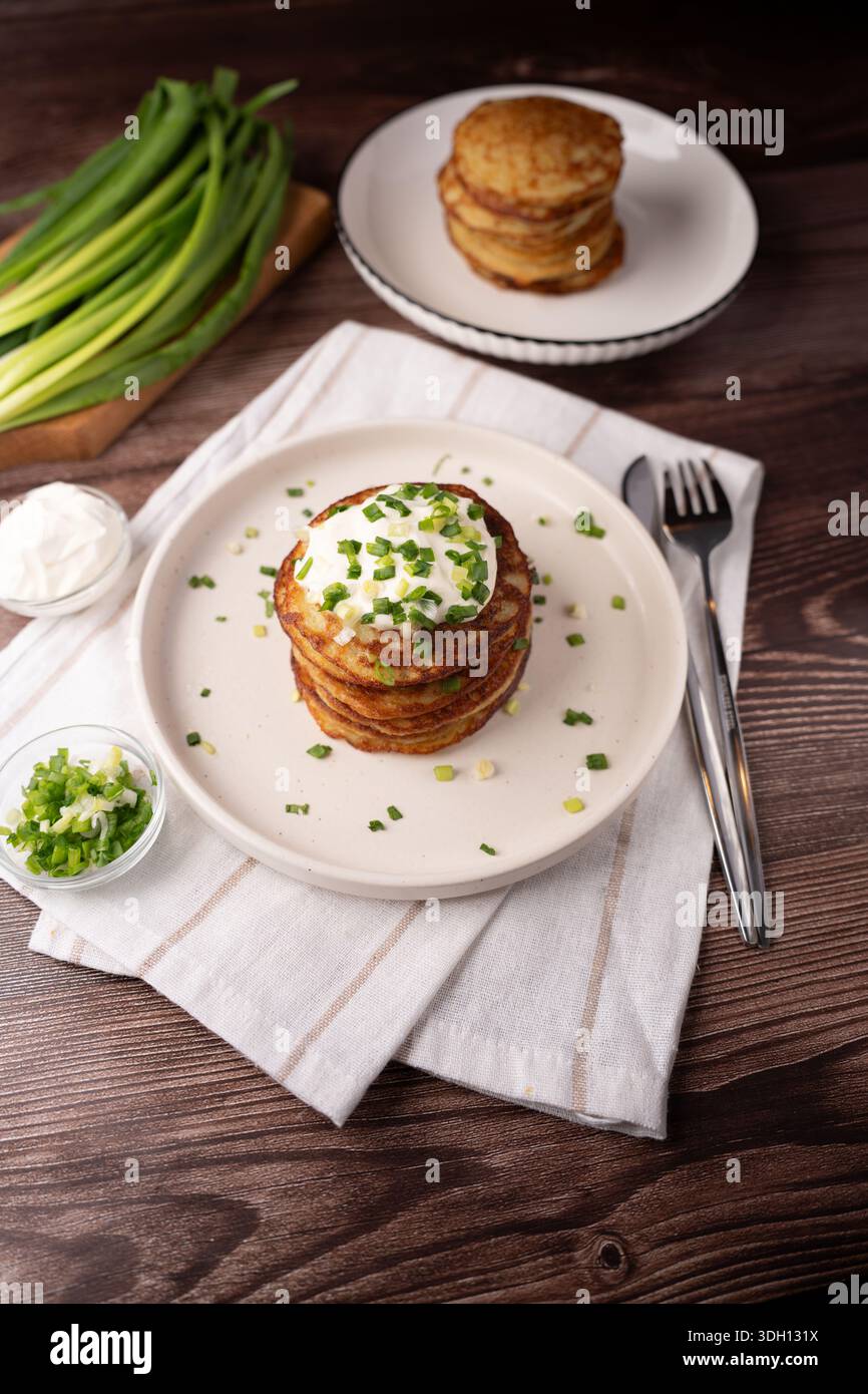 pila di frittelle di patate dorate (latkes) ricoperte di panna acida e scalpelli tritati su un piatto bianco, adagiate su legno rustico con tovagliolo di lino, ciotole e. Foto Stock