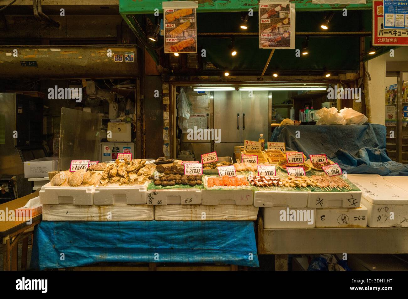 Osaka, Giappone - 7 novembre 2023: Un banco di pesce al mercato di Kuromon Ichiba espone vassoi di crostacei assortiti, ostriche, ricci di mare e spiedini di pesce con cartelli dei prezzi scritti a mano in yen giapponesi, posti su scatole di polistirolo davanti ad un banco chiuso. Foto Stock