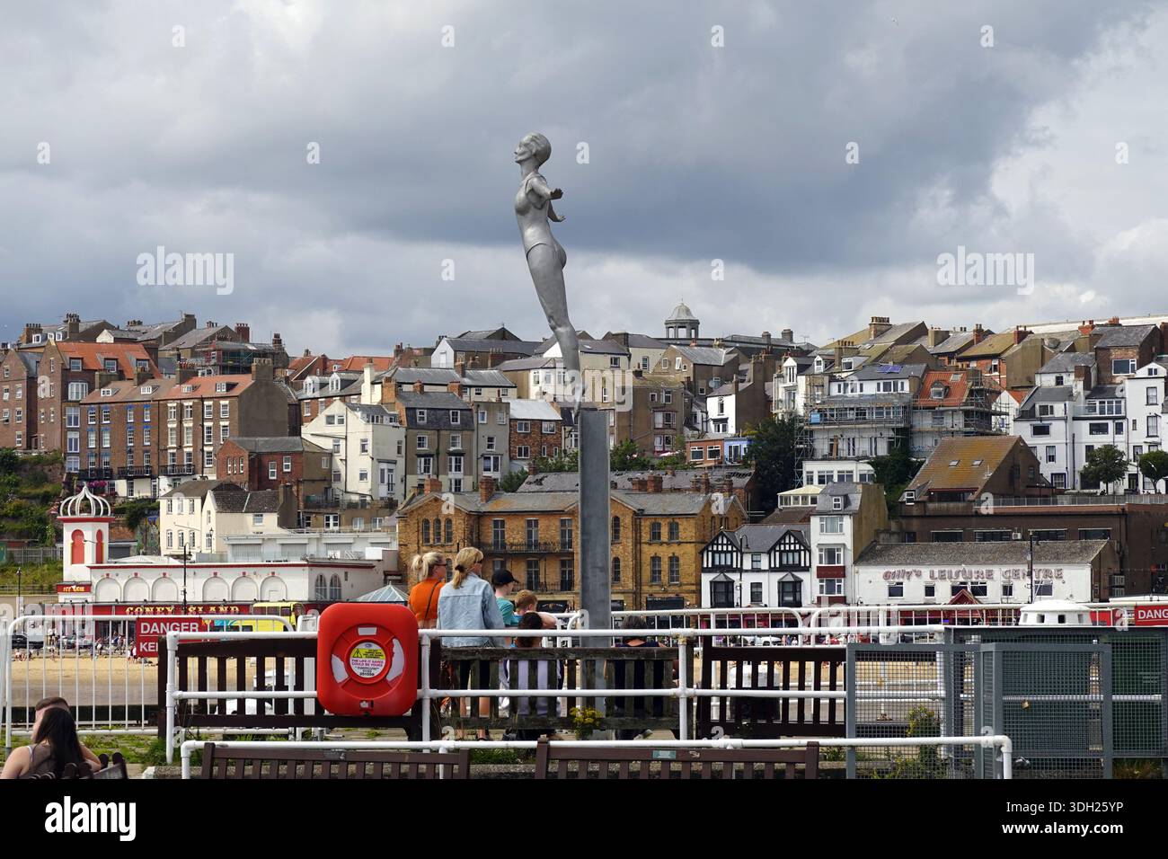 Persone sedute intorno alla statua di Diving Belle sul molo di Vincent al porto con edifici di città sul mare sullo sfondo - Scarborough, Yorkshire, Regno Unito Foto Stock