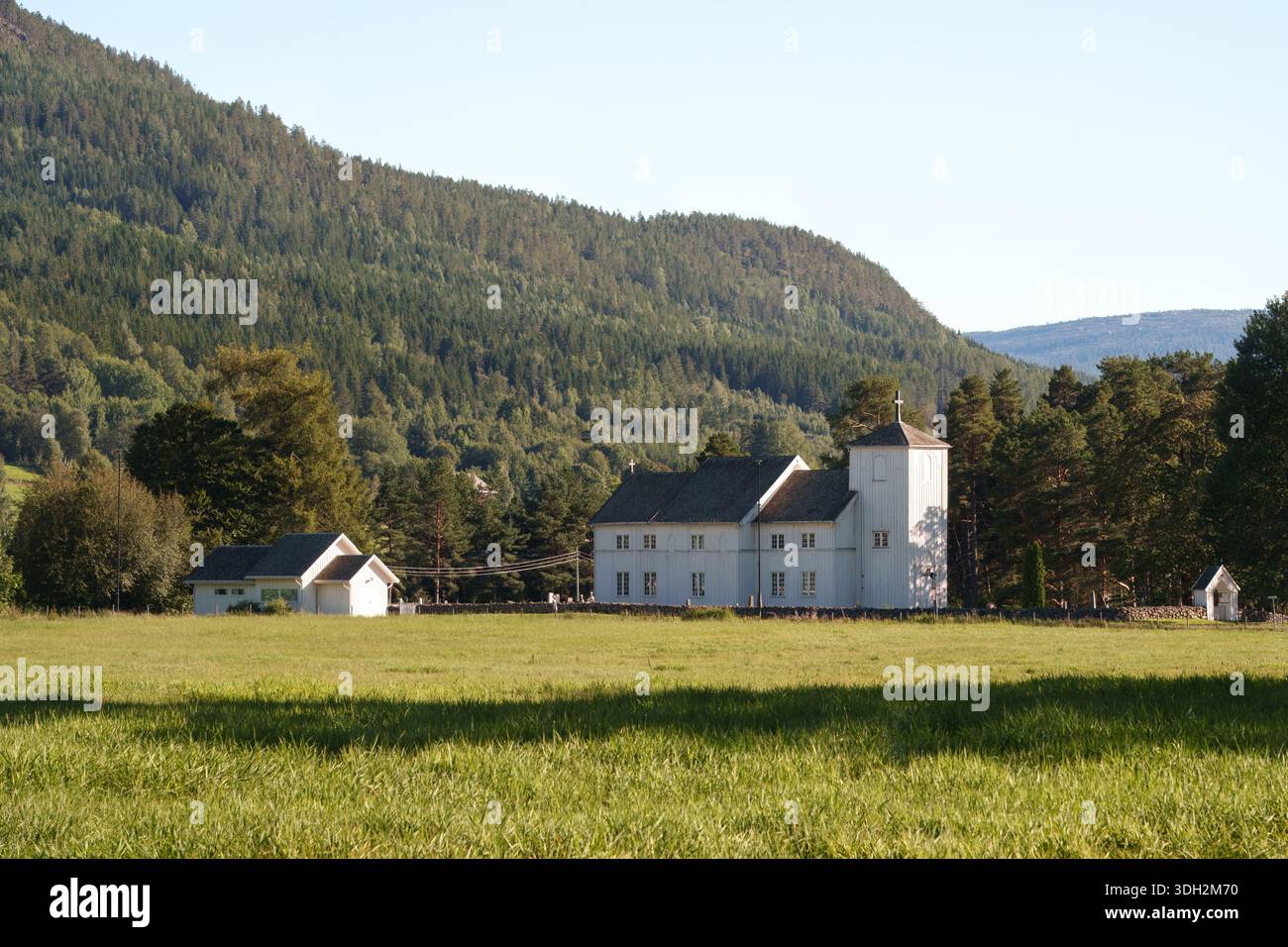 Moderna nostalgia quiete con chiesa in legno bianco adagiata su prati aperti e colline boscose, paesaggio rurale nordico limpido che evoca per lui la memoria culturale Foto Stock