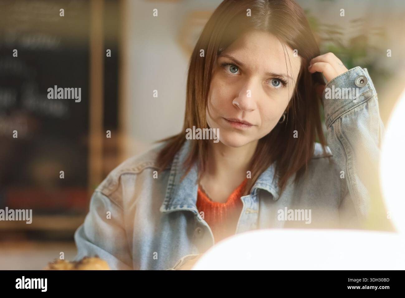 La giovane donna in giacca casual in denim appare contemplativa e distante mentre tocca i capelli in un accogliente caffè con soffice bagliore al neon Foto Stock