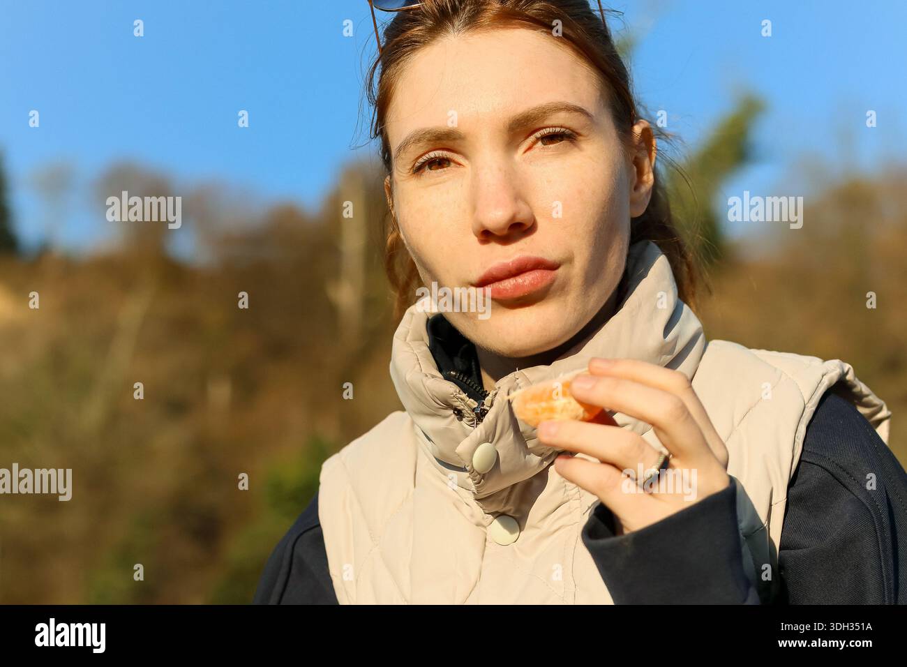 Donna che morde nel succoso segmento di mandarino all'aperto vicino al fiume calmo, occhi chiusi per divertimento Foto Stock