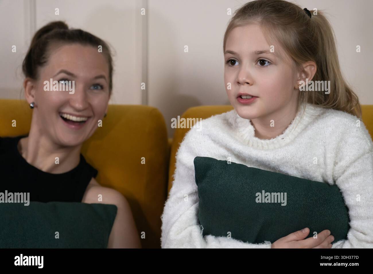 Mamma e figlia felici che si rilassano su un divano giallo senape a casa, condividendo un momento di gioia e impegnandosi in una conversazione mentre trascorrono del tempo di qualità Foto Stock