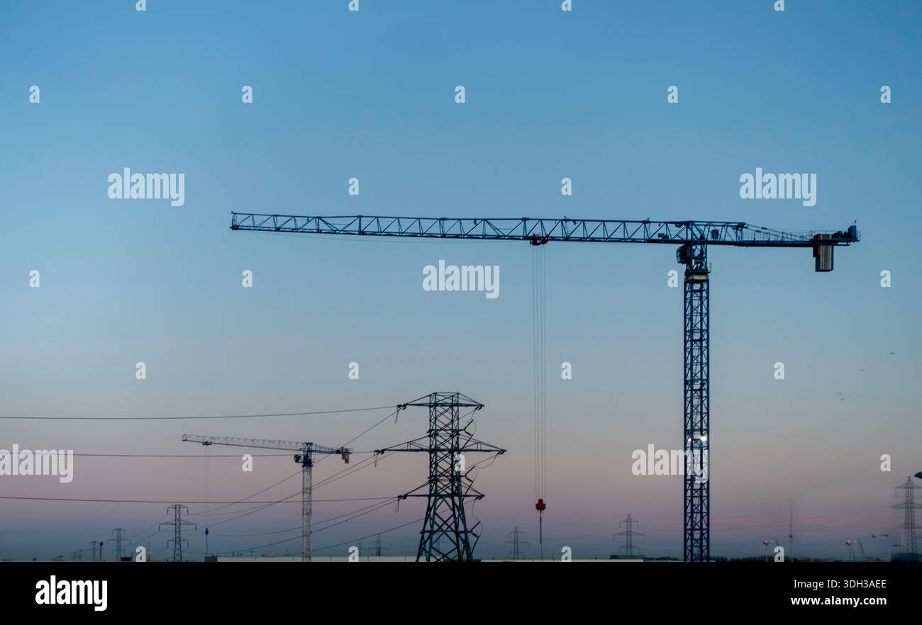 Gru industriali e tralicci elettrici si stagliano contro un cielo crepuscolo al tramonto. Infrastrutture e attrezzature per l'edilizia in un paesaggio crepuscolo. Foto Stock