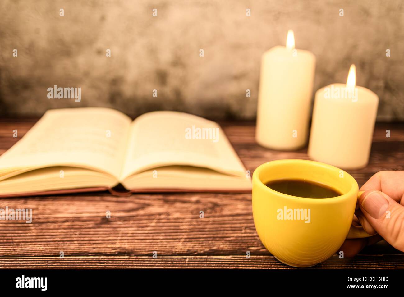 Mano che tiene una tazza di caffè su un tavolo in legno rustico, con libro aperto e candele a fuoco morbido. Atmosfera accogliente con spazio fotocopie. Foto Stock