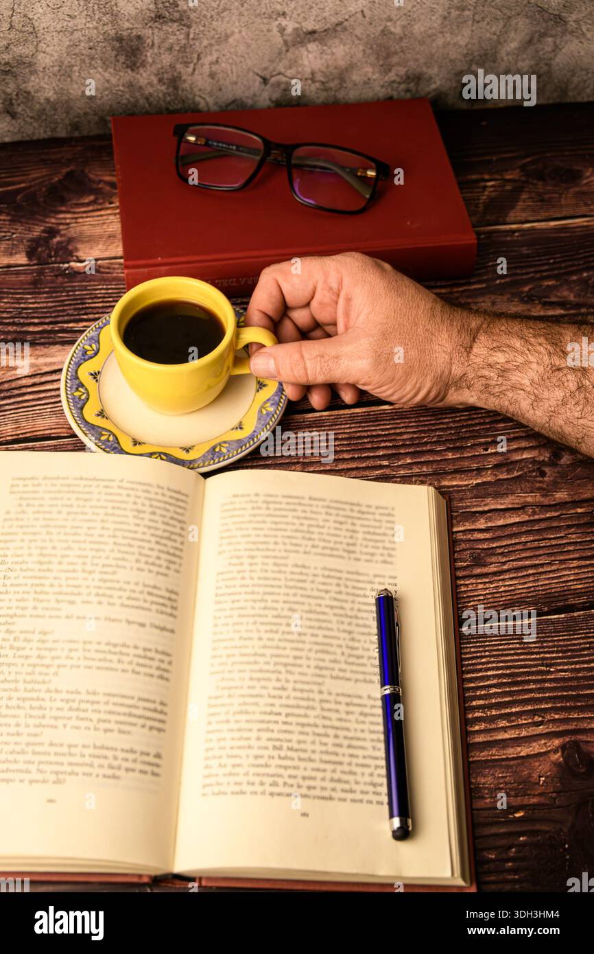 Vista dall'alto di un libro aperto con una penna e una mano che tiene il caffè su una scrivania rustica in legno. Studio, scrittura e concetto di produttività. Foto Stock