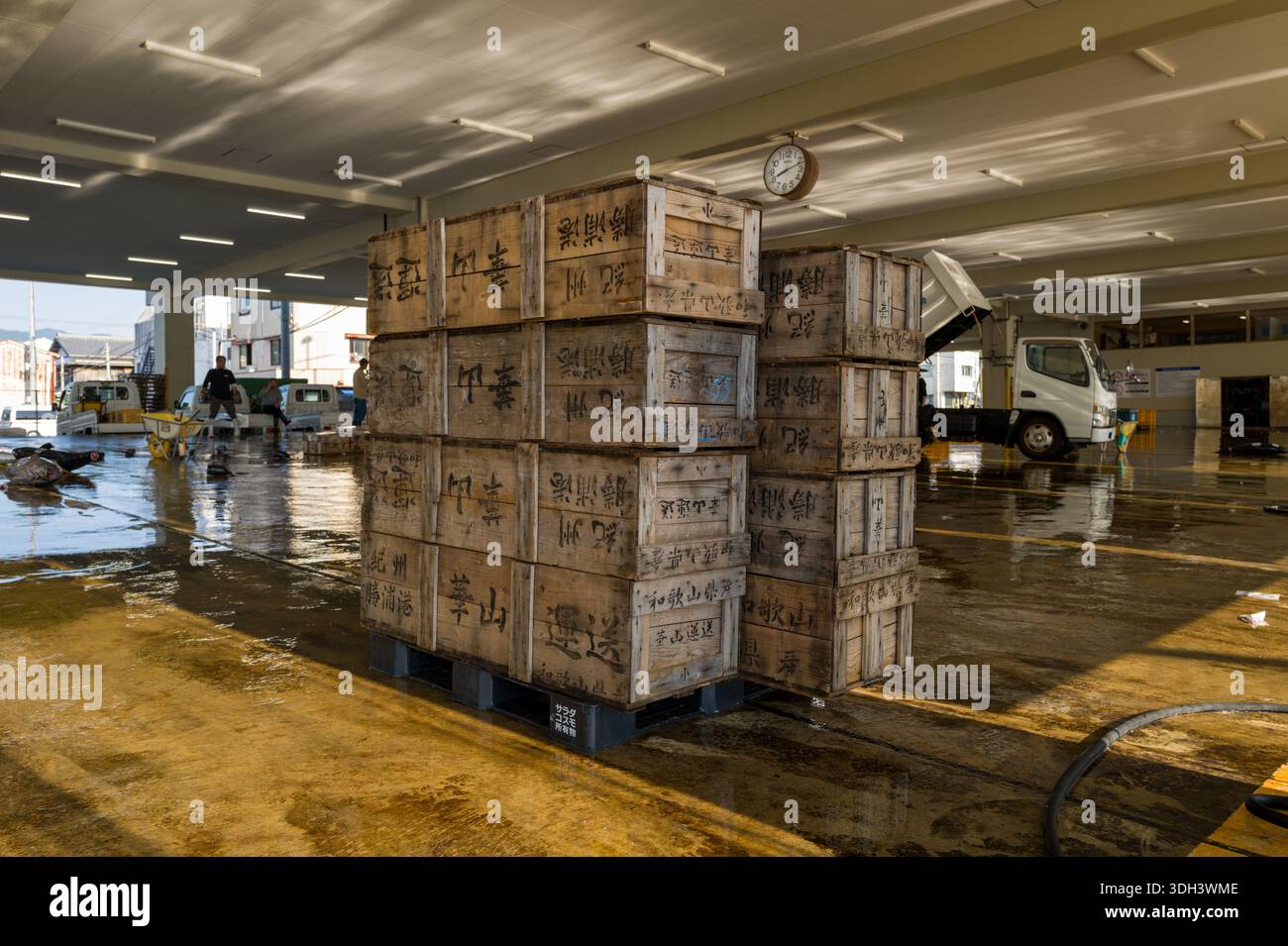 Nachikatsuura, Giappone - 27 ottobre 2023: Pile di casse di legno con scritta giapponese sono disposte su pallet all'interno di un mercato del pesce coperto, con un orologio sul soffitto, pavimenti bagnati e lavoratori e veicoli visibili sullo sfondo. Foto Stock