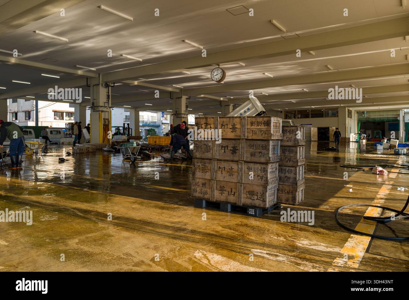 Nachikatsuura, Giappone - 27 ottobre 2023: I lavoratori gestiscono attrezzature e casse di legno con scritta giapponese all'interno di un mercato del pesce coperto, con orologio, pavimento bagnato e vari strumenti di mercato visibili. Foto Stock