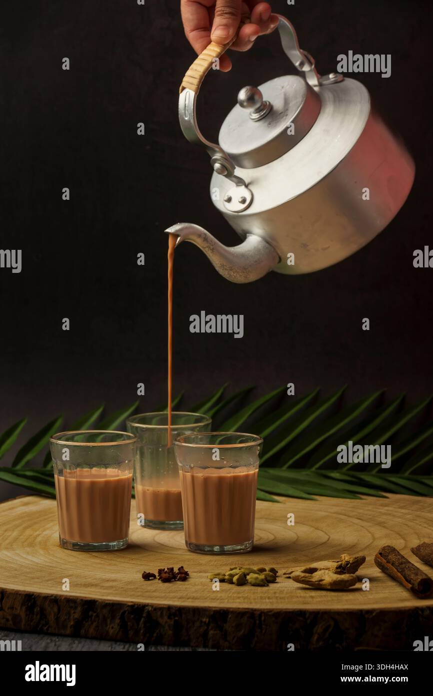 Una persona sta versando tè masala in tre tazze chai di vetro. Le tazze sono su un tavolo di legno. Tagliare il chai che viene versato dal bollitore del tè. Foto Stock