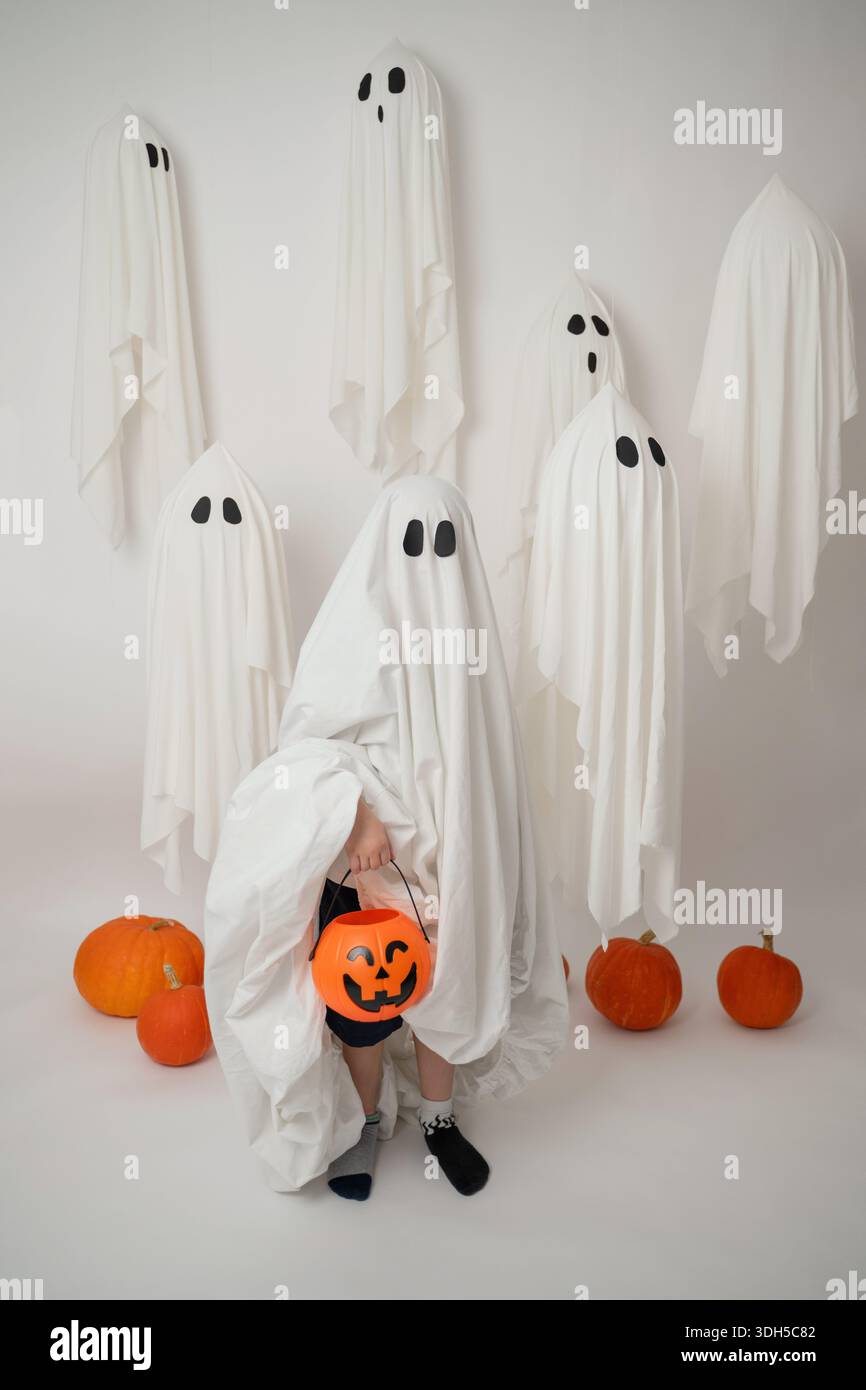 Ritratto natalizio di Halloween con un bambino in costume fantasma che regge un secchio di zucca. Intorno a loro ci sono spaventose lenzuola fantasma fluttuanti e vibra Foto Stock