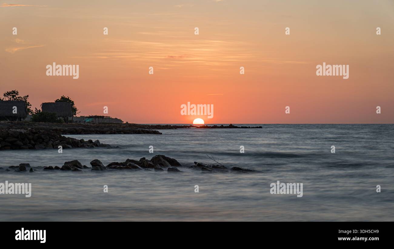 Un vivace tramonto dipinge la spiaggia dei Caraibi in tonalità calde. Il sole tramonta sulle acque calme, creando un'atmosfera serena con sagome di rocce e dist Foto Stock