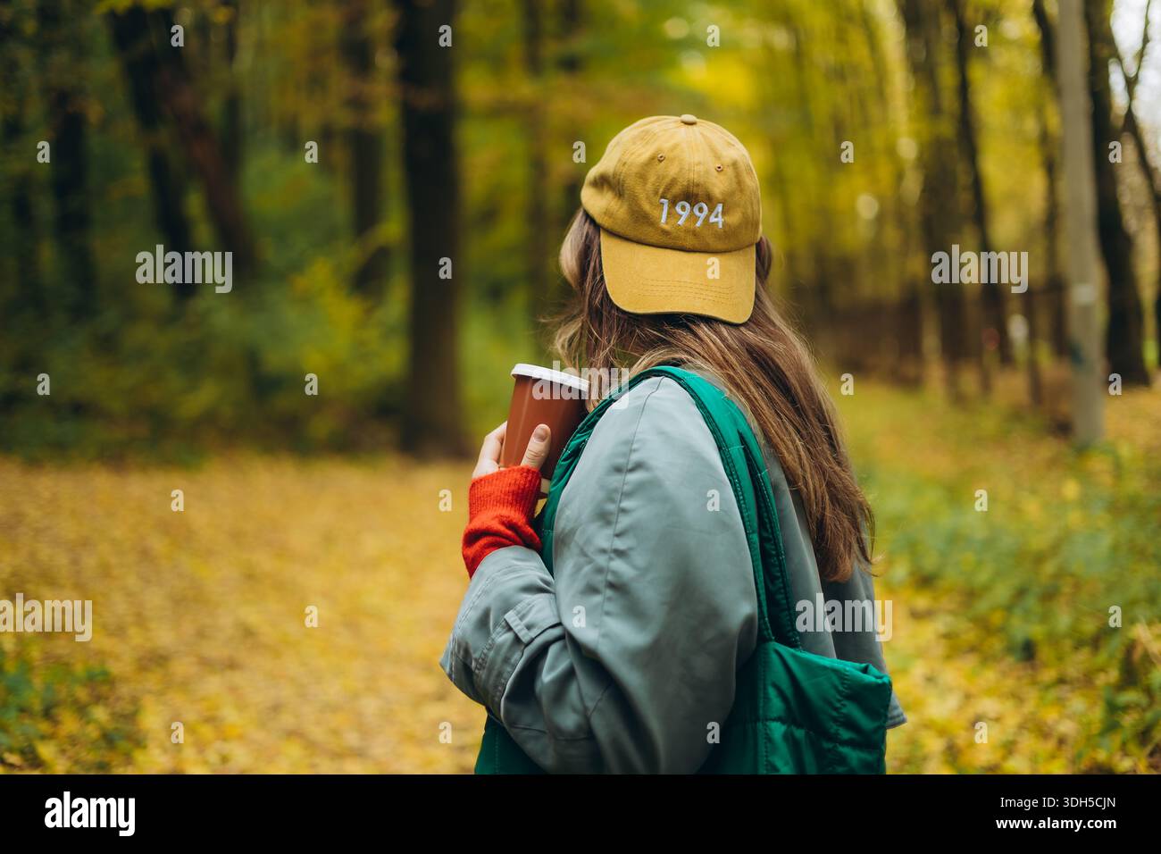 Una ragazza gode di una tranquilla passeggiata autunnale attraverso una lussureggiante foresta, tenendo in mano una tazza di caffè caldo. Indossa un outfit casual e un cappellino, che abbracciano il vivace design Foto Stock
