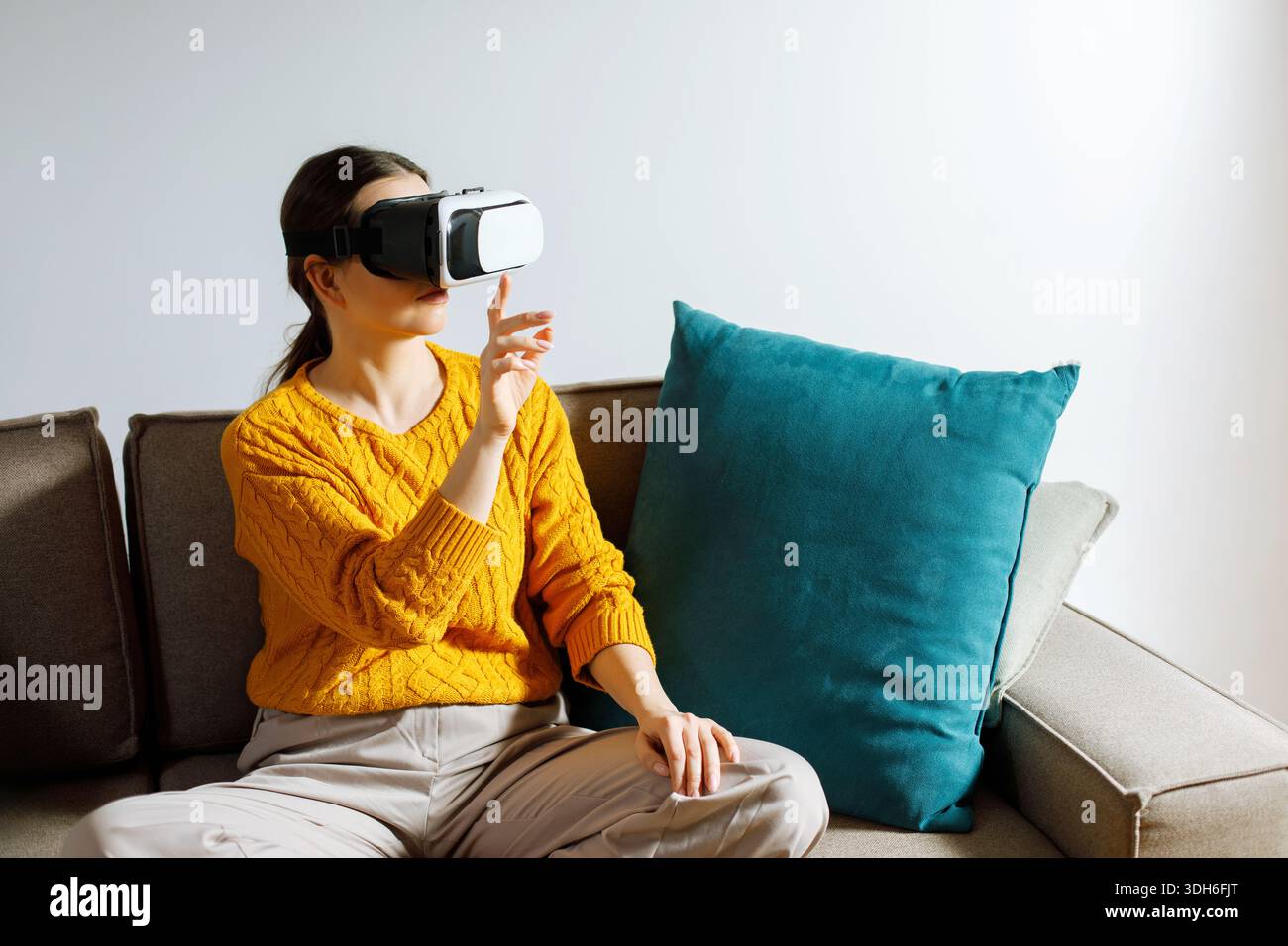Una donna siede tranquillamente su un divano mentre indossa un visore per la realtà virtuale. Sembra concentrata e impegnata con l'esperienza virtuale nel suo Roo vivente Foto Stock