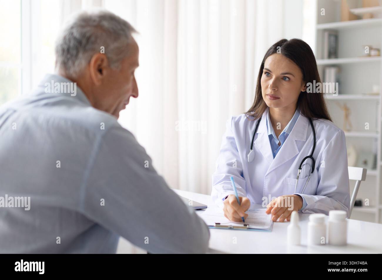 Donna medico consulenza paziente di sesso maschile senior alla scrivania e scrittura note mediche Foto Stock
