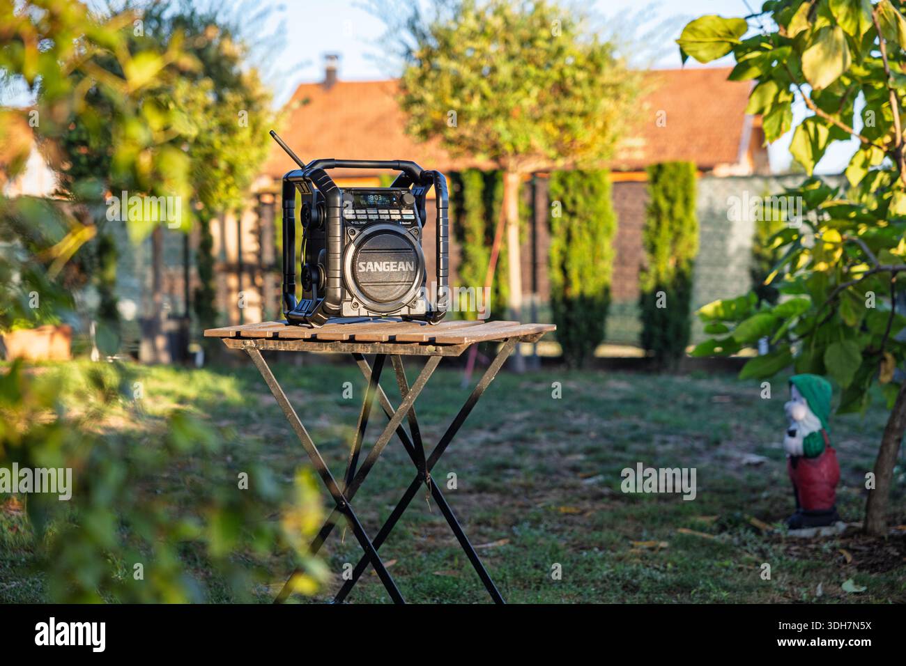 VINKOVCI, CROAZIA, 09.19.2025: Una robusta radio portatile poggia su un rustico tavolo in legno in un giardino soleggiato, con una stravagante statua di gnomi nelle vicinanze e vibra Foto Stock