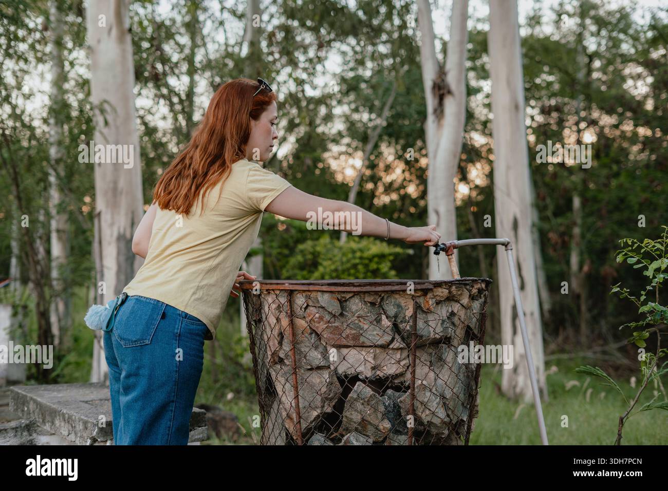 Giovane donna dai capelli rossi che si avvicina a un cestino di legna da ardere pieno di tronchi in un parco, concetto di preparazione all'aperto. Foto Stock
