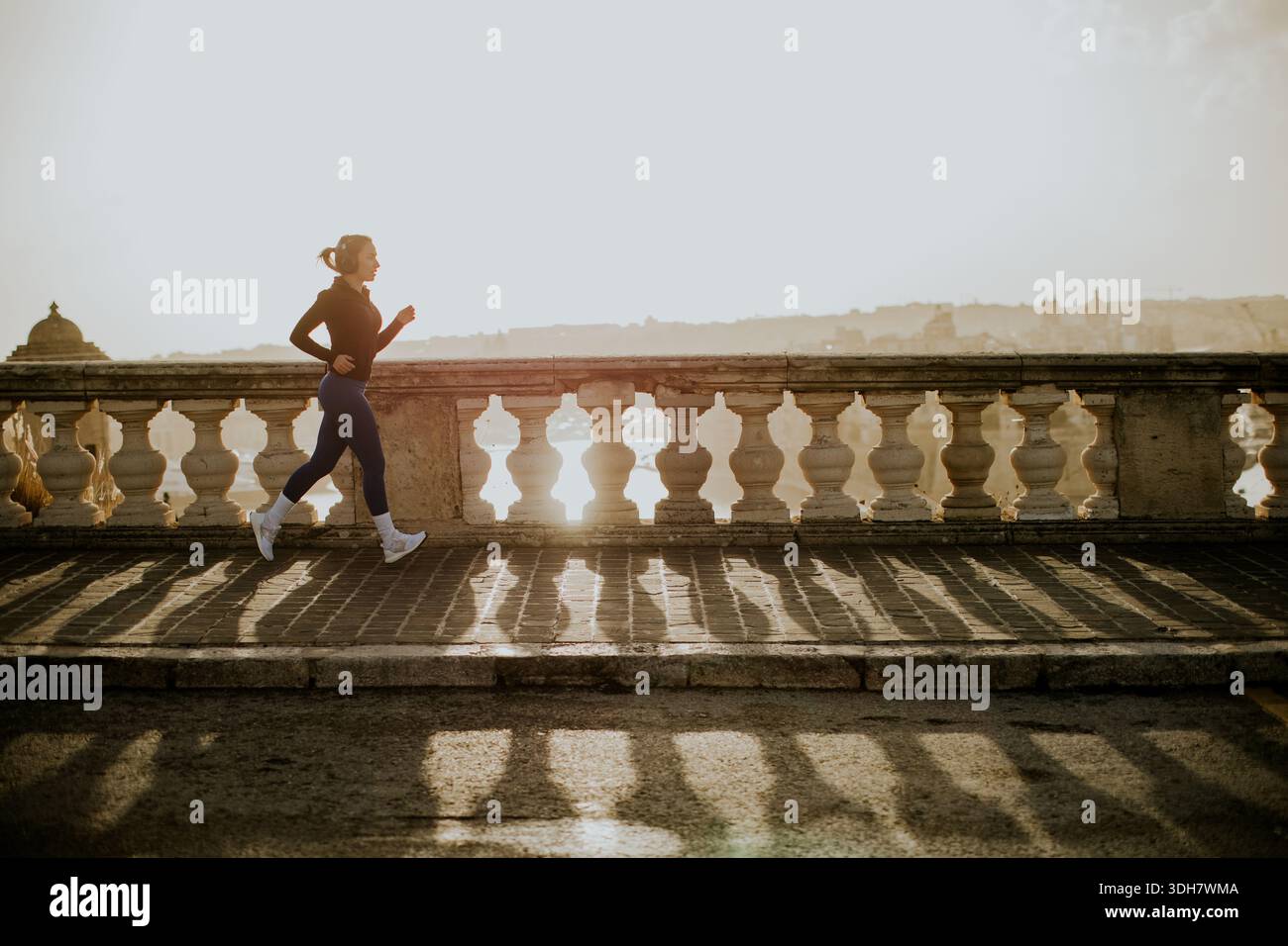 Una donna fa jogging su una passerella vicino al mare a Malta durante il tramonto. La luce del sole brilla, creando lunghe ombre e un'atmosfera calda intorno a lei Foto Stock