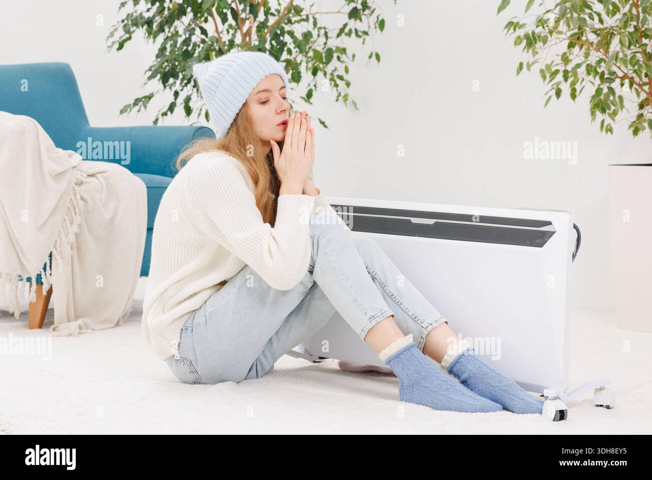 Una giovane donna in abiti caldi siede sul pavimento con un riscaldatore elettrico portatile in una casa fredda durante l'inverno, risparmiando energia Foto Stock