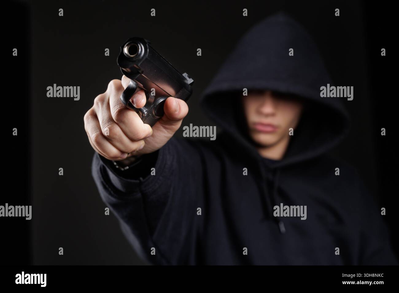 Persona con cappuccio che punta la pistola alla telecamera in una scena buia che rappresenta un pericolo di rapina a mano armata e un concetto di rischio per la sicurezza Foto Stock