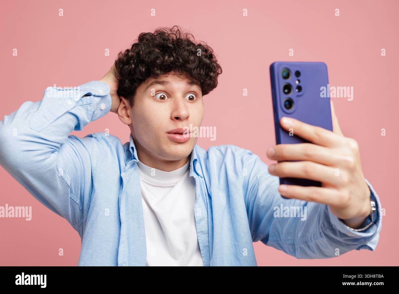 Giovane scioccato dalla notifica inaspettata dello smartphone mentre controlla lo schermo isolato su sfondo rosa Foto Stock
