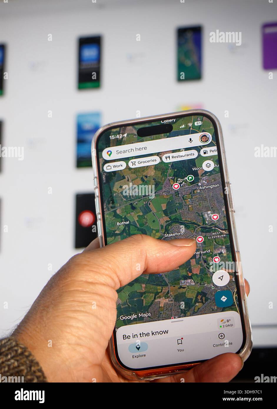 Persona che tiene in mano lo smartphone utilizzando l'app di navigazione Google Maps mentre gli schermi digitali sono visibili sfocati sullo sfondo Foto Stock