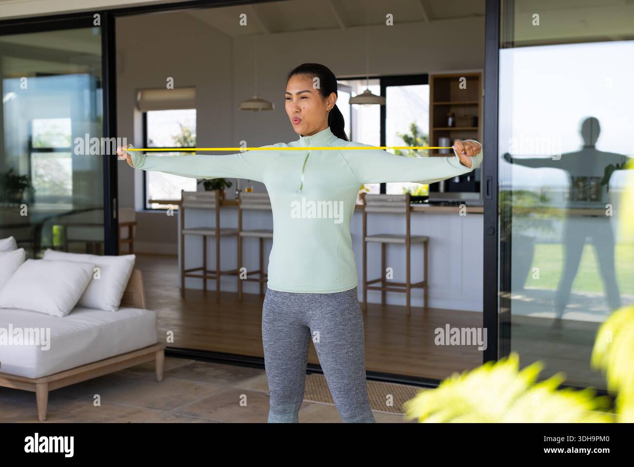 Donna asiatica di mezza età che tiene la fascia gialla che si estende sul petto sul patio indossando un pullover Foto Stock