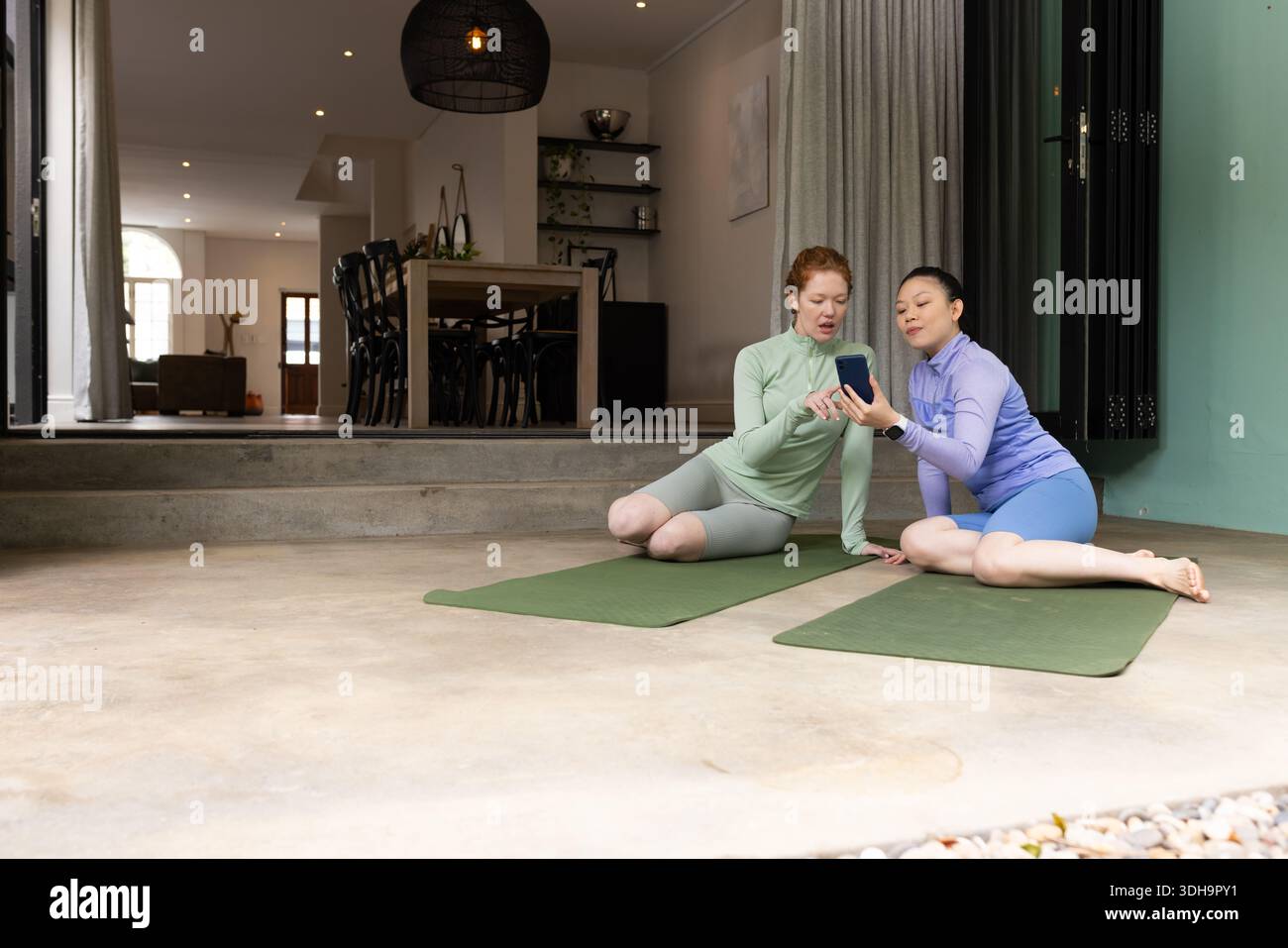 Diverse amiche che siedono su tappetini verdi da yoga su top atletiche condividendo telefono e spazio per copiare Foto Stock