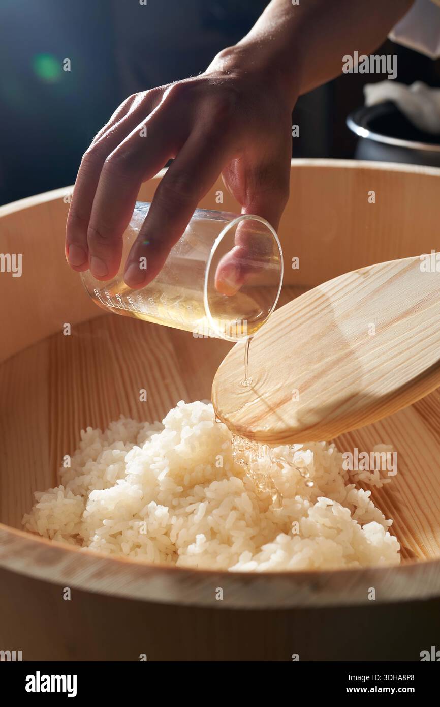 Lo chef versa aceto di riso stagionato in una tradizionale ciotola di hangiri in legno con riso bianco al vapore per la preparazione del sushi. Foto Stock