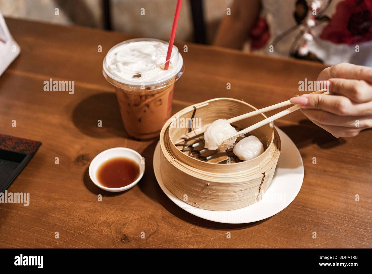 A mano con bacchette che raccolgono dim sum nel piroscafo di bambù e tè tailandese freddo su un tavolo di legno Foto Stock
