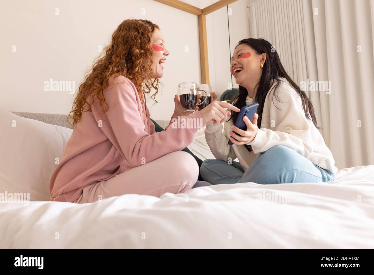 Diverse donne e amici ridono, tenendo vino e smartphone a letto in abiti da salotto, cerotti in idrogel Foto Stock
