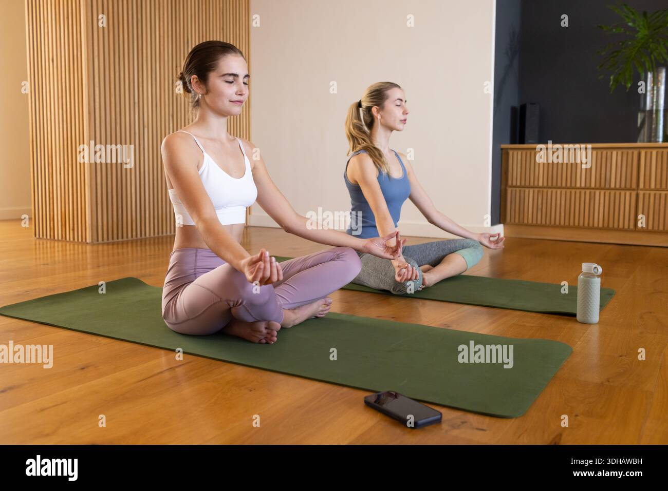Amiche che meditano sui tappetini verdi in studio, indossano abbigliamento sportivo, telefono e bottiglia d'acqua nelle vicinanze Foto Stock